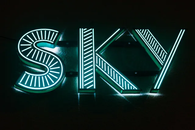 Illuminated neon sign displaying 'SKY' in a dark setting, glowing blue-green. Profitable sign shop services: costing one channel letter job